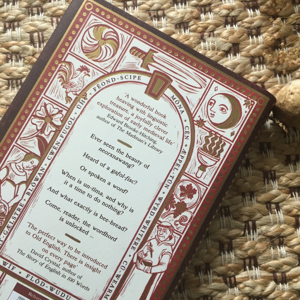 The back cover of a book against a background of woven textiles. The cover background is white with a gold and red border with decorative gold dots. The text within an arched window shape are quotations from Edward Brooke-Hitching and David Crystal. ‘A blurb for the book says: ‘Ever seen the beauty of neorxnawang? Heard of a gafol-fisc? Or spoken a word? When is un-tima, and why is it a time to do nothing? And what exactly is bee-bread? Come, reader, the wordhord is unlocked… A border of Old English words surrounds the window: blowan, cwen-fugol, uht, feond-scipe, mona, cen, æppel-tun, wyrd-writere, cu-wearm, wif, and flod-wudu. The area surrounding the Old English word border has wood-cut style illustrations in red and gold, each in its own compartment but overlapping slightly: a flower, a rooster, the sun, a sword, the moon and stars, a torch, grapes on a vine, and a monk writing in a book.