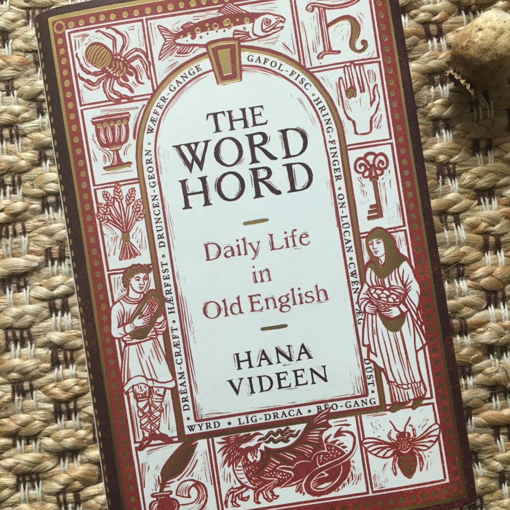 A book, The Wordhord: Daily Life in Old English, by Hana Videen, against a background of woven textiles. The cover background is white with a gold and red border with decorative gold dots. The title and author’s name are written inside an arched window shape, with a border of Old English words surrounding it: wæfre-gange, gafol-fisc, hring-finger, on-lucan, cwen, æg, dust, beo-gang, lig-draca, wyrd, dream-cræft, hærfest, and druncen-georn. The area surrounding the Old English word border has wood-cut style illustrations in red and gold, each in its own compartment but overlapping slightly: a spider, a fish, the letter h in a calligraphic style, a hand with a ring, a key, a woman carrying a basket of eggs, a bee, a dragon, an ink pot and quill with scroll of paper, a man playing a harp, a bundle of wheat, and a goblet.