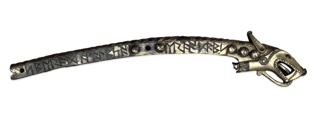 A slightly curved silver-gilt fitting with runes inscribed along the side; one end is decorated with an intricate three-dimensional animal head with long ears, fangs and a tongue curling out so far out of its mouth that it touches its throat.