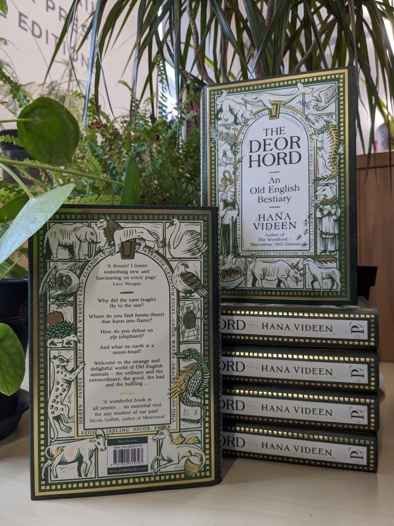 A stack of the book “The Deorhord: An Old English Bestiary” by Hana Videen, with one book on top of the stack showing the front cover and another book leaning against it showing the back cover, with plants in the background. The cover is green and gold, decorated with various animals and strange creatures in wood-cut style illustrations, and the frame around the text contains Old English words.