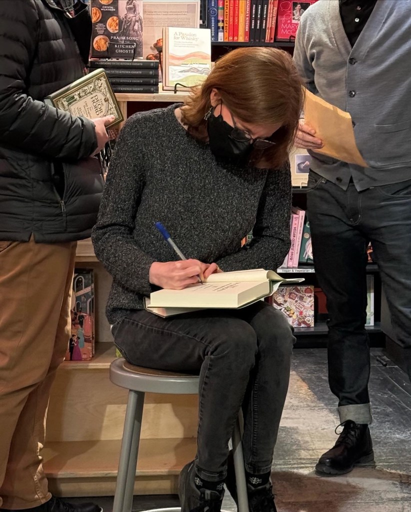 Hana Videen sitting on a stool and signing a copy of her book.