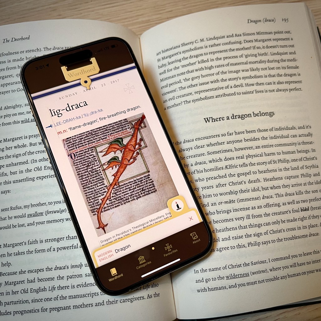 An iPhone showing the Wordhord app with the word dragon in a search field and the word lig-draca displayed above with the definition flame-dragon or fire-breathing dragon and a medieval manuscript image of a red dragon. The phone sits on a hardcover copy of The Deorhord open to a page with a heading “Where a dragon belongs”.
