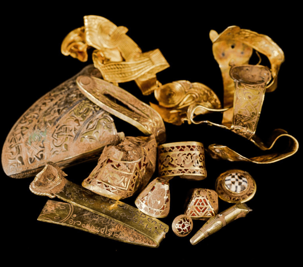 A pile of worn and damaged gold items, including part of a cheek piece, a hilt with an intertwining pattern, mounts and studs with inlaid patterns, and a folded band with a Latin inscription.