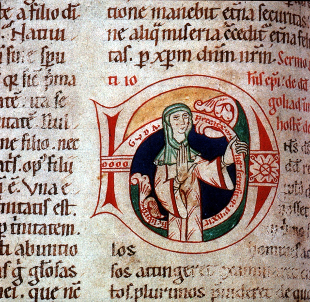 A historiated initial in a medieval manuscript depicting a nun raising her right hand before her and gripping a Latin text band that curves around her in her left hand.